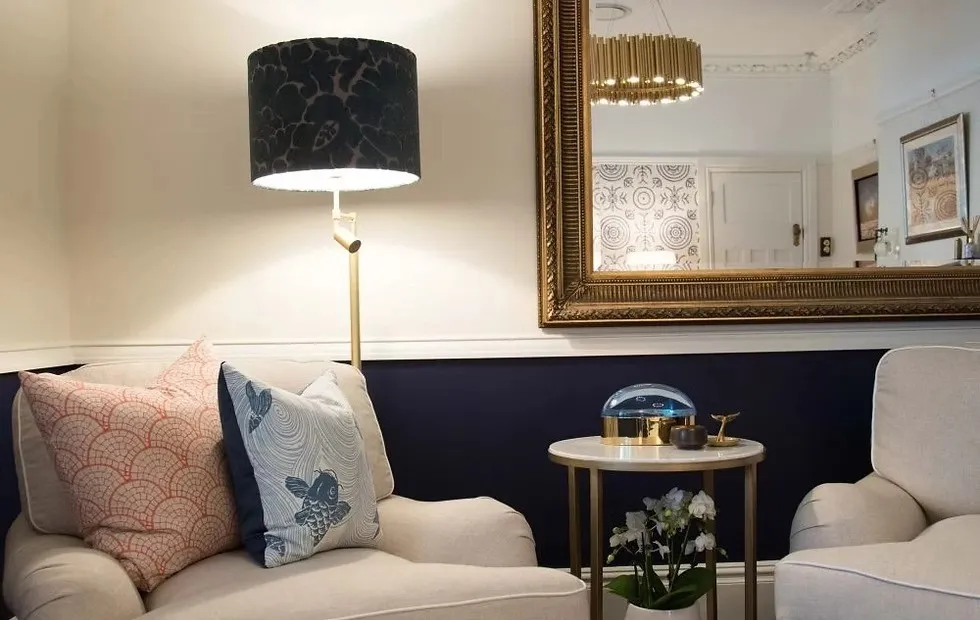Cozy seating area with two beige armchairs, patterned accent pillows, a small round side table with decorative items, a tall floor lamp with a black shade, and a large ornate gold-framed mirror on a navy and white wall.